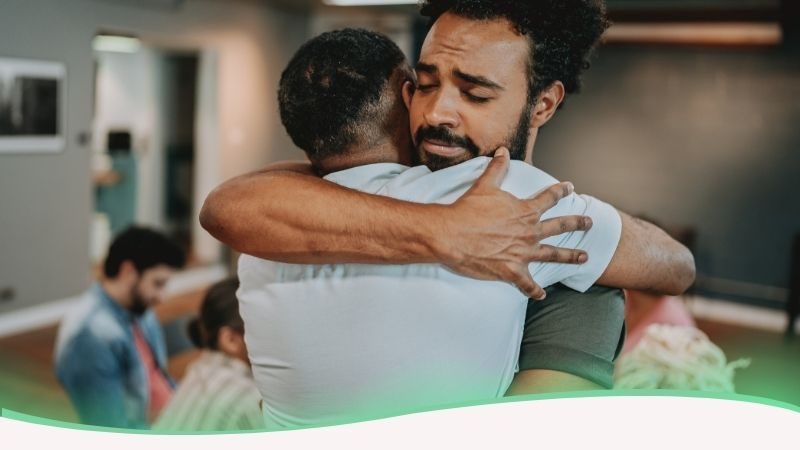 Uma imagem emocionante e reconfortante que mostra dois homens em um abraço fraterno e sincero. Um deles apoia a cabeça no ombro do outro, revelando uma expressão de vulnerabilidade e alívio, enquanto o amigo o segura com firmeza, transmitindo segurança e compreensão. A linguagem corporal de ambos demonstra uma conexão profunda e um suporte genuíno. A imagem ilustra o tema "A possibilidade de lidar com a dor de uma paixão existe dentro de cada um": simboliza que a força para superar um sofrimento amoroso também se encontra na nossa capacidade de pedir e receber ajuda. O abraço representa o porto seguro onde a dor pode ser expressa sem julgamentos, reforçando que o apoio humano é uma das ferramentas mais poderosas para transformar a angústia em aceitação e recomeço