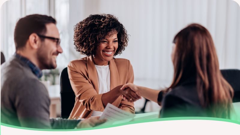 Fotografia de uma sala de reuniões onde um casal de selecionadores (um homem e uma mulher) conduz uma entrevista com uma candidata. Os entrevistadores estão sentados à mesa com laptops e documentos, demonstrando atenção e profissionalismo. A moça está posicionada de frente para eles, transmitindo uma postura aberta e engajada. A imagem ilustra o cenário real das perguntas mais feitas pelo selecionador, destacando a interação profissional e a importância da preparação para diferentes formatos de avaliação