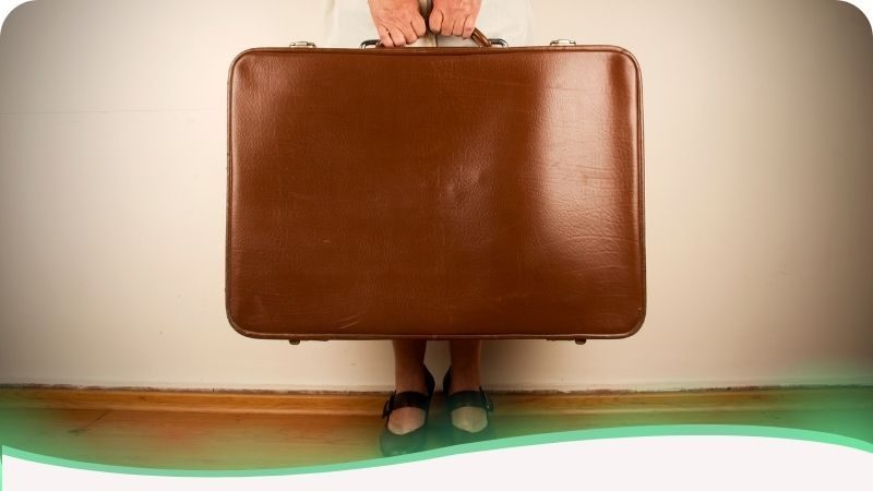 Uma imagem enquadrada de frente que apresenta uma mala de viagem marrom de proporções gigantescas, ocupando quase todo o plano. Atrás dela, nota-se a presença de uma mulher, da qual apenas as mãos são visíveis — segurando as alças com esforço — e os pés aparecem na parte inferior, sustentando o peso. A figura humana é quase totalmente eclipsada pelo volume da bagagem. A imagem ilustra o subtítulo sobre não se desgastar em um relacionamento abusivo: simboliza o peso desproporcional que uma das partes carrega sozinha na tentativa de manter a relação. Representa o esgotamento físico e mental de quem transporta fardos emocionais excessivos, evidenciando como a pessoa acaba desaparecendo atrás das demandas e problemas do outro