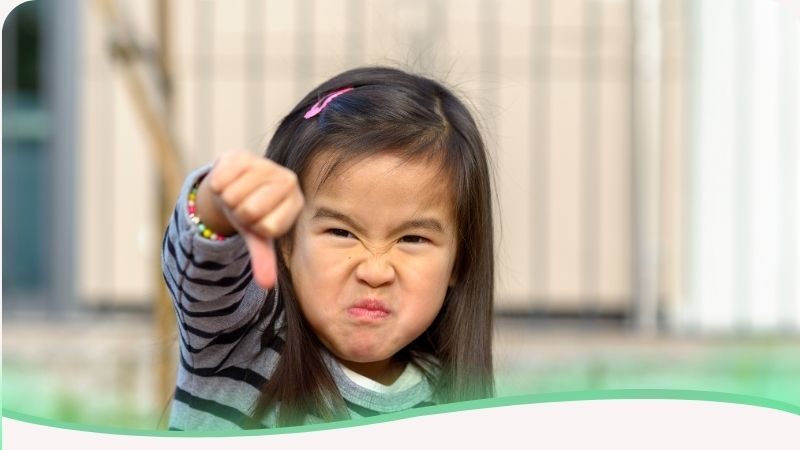 Criança asiática fazendo uma careta de birra e gesticulando com o polegar para baixo (descurtir), representando a resistência em aceitar ordens e o comportamento opositor típico da Síndrome do Imperador