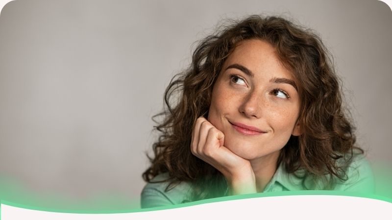 Um retrato encantador e reflexivo de uma jovem mulher branca com cabelos cacheados volumosos. Ela está com a mão apoiada delicadamente no queixo e o olhar direcionado para cima, voltado para o título do texto, com um leve e sereno sorriso nos lábios. Sua expressão transmite uma mistura de curiosidade, satisfação e esperança. A imagem ilustra o tema "A teoria da motivação de Maslow": simboliza a jornada humana através da pirâmide das necessidades. O olhar para cima representa a busca pelos níveis mais altos da teoria, como a estima e a autorrealização. O sorriso sutil reflete a motivação interna de quem compreende suas próprias faltas e potências, mostrando que a teoria de Maslow não é apenas um conceito, mas um mapa vivo para o crescimento pessoal e a conquista da plenitude